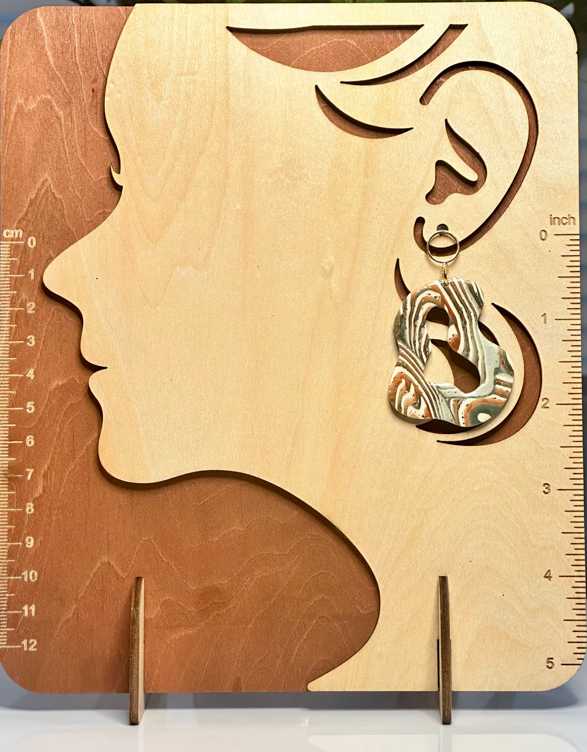 A single dangling earring displayed on a wooden silhouette stand shaped like a woman's head in profile, with ruler markings along the left and right edges. The earring has an abstract marbled pattern in orange, brown, black, and white, hanging from a gold-toned ring.